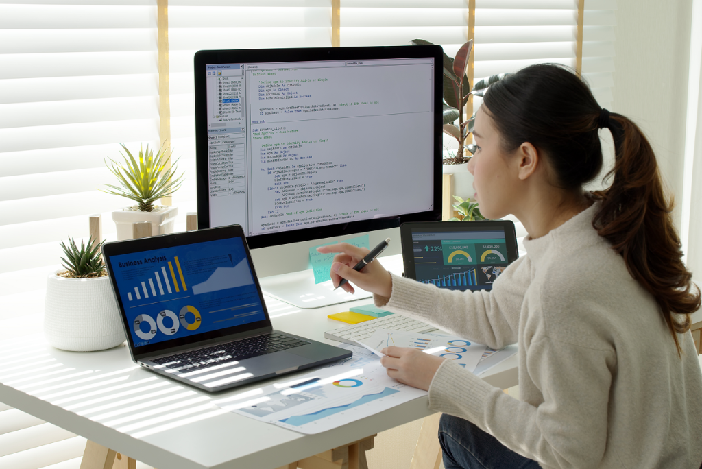 Female seated around a desk with a desktop monitor, laptop. tablet, and paper showing charts